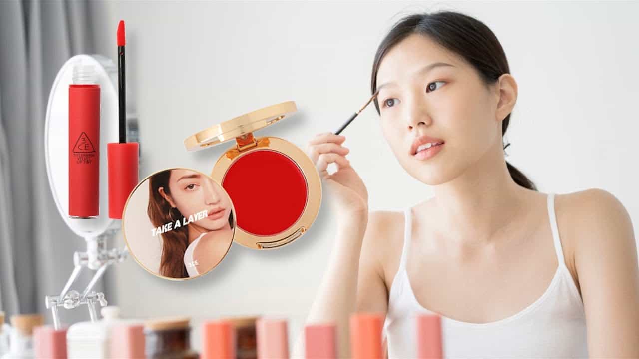 A woman applies red lipstick, surrounded by cosmetics and a mirror, smiling gently.