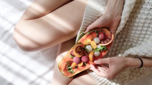 A person holding a halved papaya filled with colorful fruits, sitting on a cozy blanket.