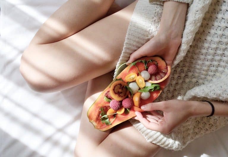 A person holding a halved papaya filled with colorful fruits, sitting on a cozy blanket.